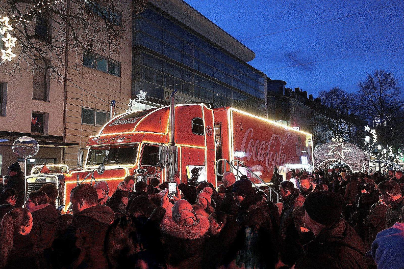 The illuminated Coca-Cola Christmas truck surrounded by a crowd taking photos at a Christmas market