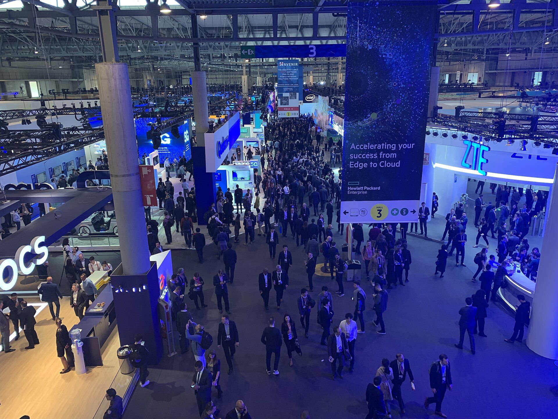 Looking down on a crowded exhibition hall at MWC Barcelona, lined with illuminated stands